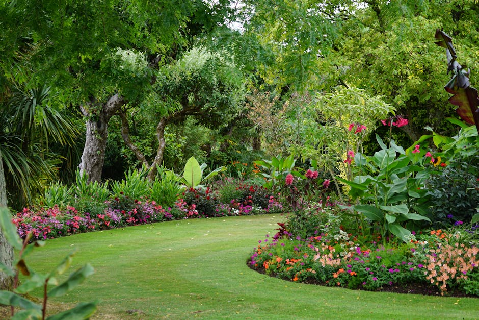 A lush, well-maintained garden featuring a curved, freshly mowed lawn bordered by vibrant flower beds with a variety of blooming plants in shades of pink, orange, and purple. Tall, leafy trees with te
