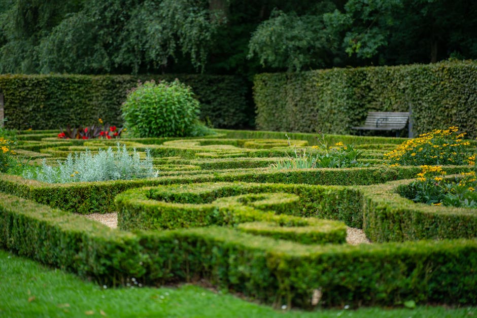 A lush, well-maintained garden featuring a curved, freshly mowed lawn bordered by vibrant flower beds with a variety of blooming plants in shades of pink, orange, and purple. Tall, leafy trees with te